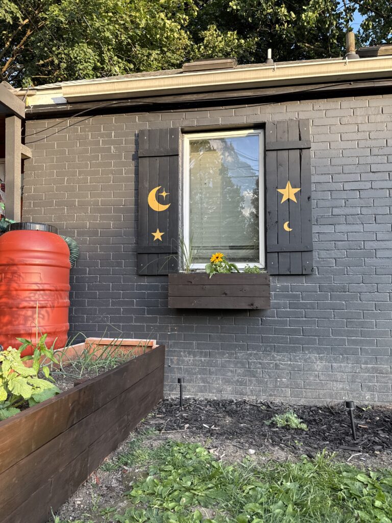 Handmade black wooden window shutters decorated with gold moons and stars beside a raised garden bed.