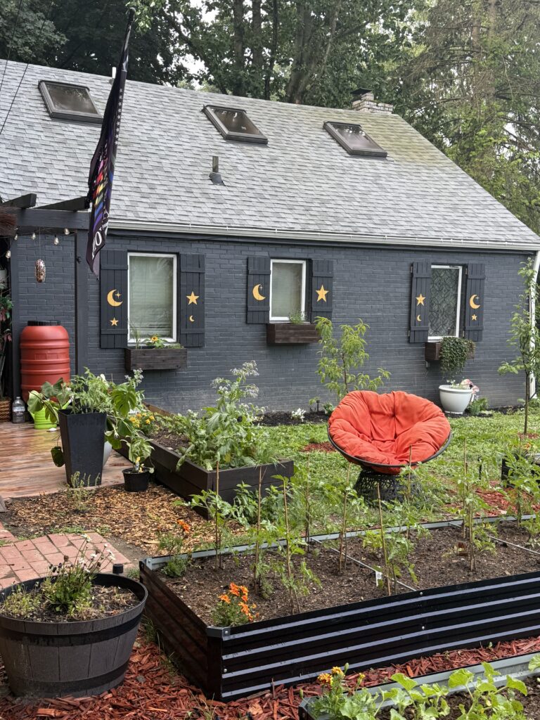 Raised vegetable garden beds beside a dark-painted cottage with handmade moon and star shutters.